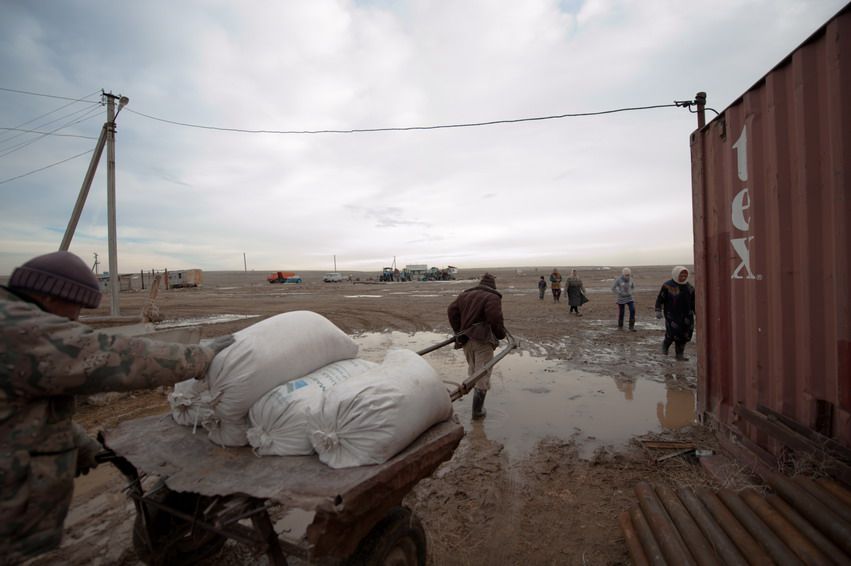 Мужчины везут мешки на тележке по грязной дороге фермы после дождя, вдалеке видны люди и степь. ферма, мешки, работа, Казахстан, верблюжье хозяйство