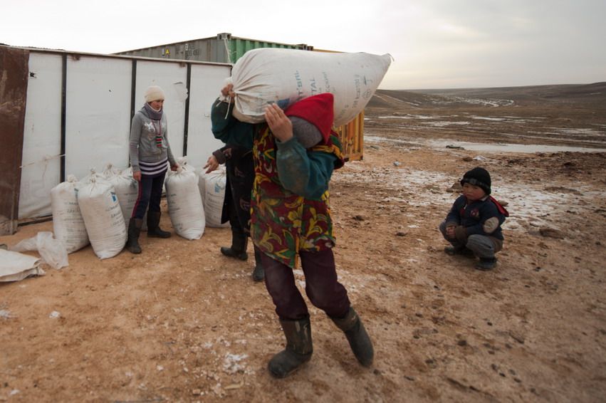 Женщина несёт тяжёлый мешок на плече, рядом стоит ребёнок и другая женщина на фоне контейнеров. Сельская жизнь, семья, труд, степь, верблюжье хозяйство