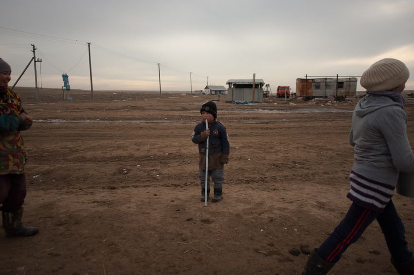Маленький мальчик стоит с палкой посреди степной дороги возле женщин и построек. ребёнок, село, Казахстан, степь, верблюжье хозяйство